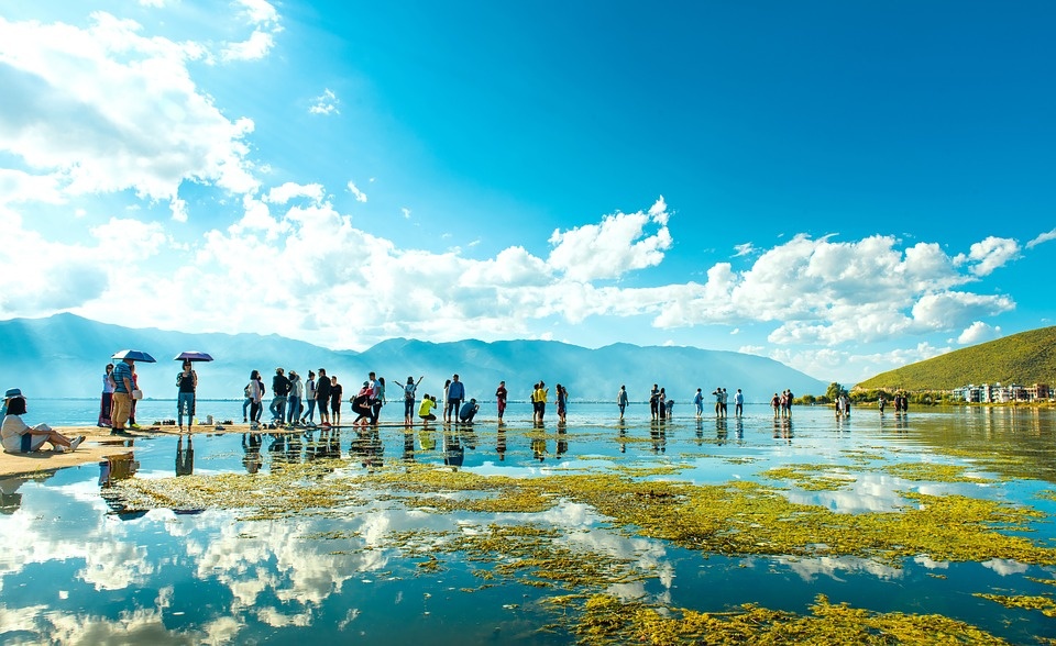 Travelers beside a shallow lake reflecting blue skies, white clouds, and distant mountains