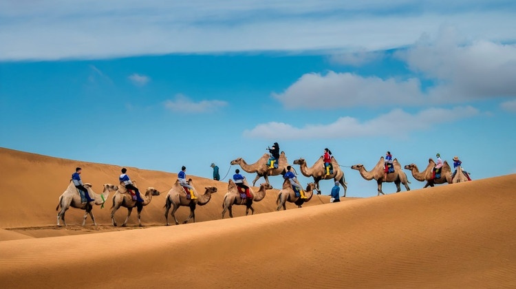 Travelers riding camels across golden desert dunes beneath a blue sky